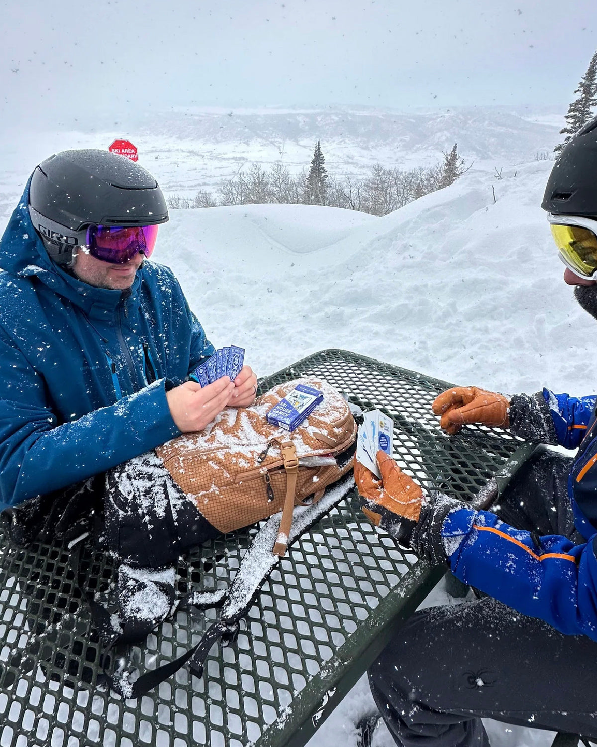 Two skiers playing cards on a snowy picnic table at a ski resort