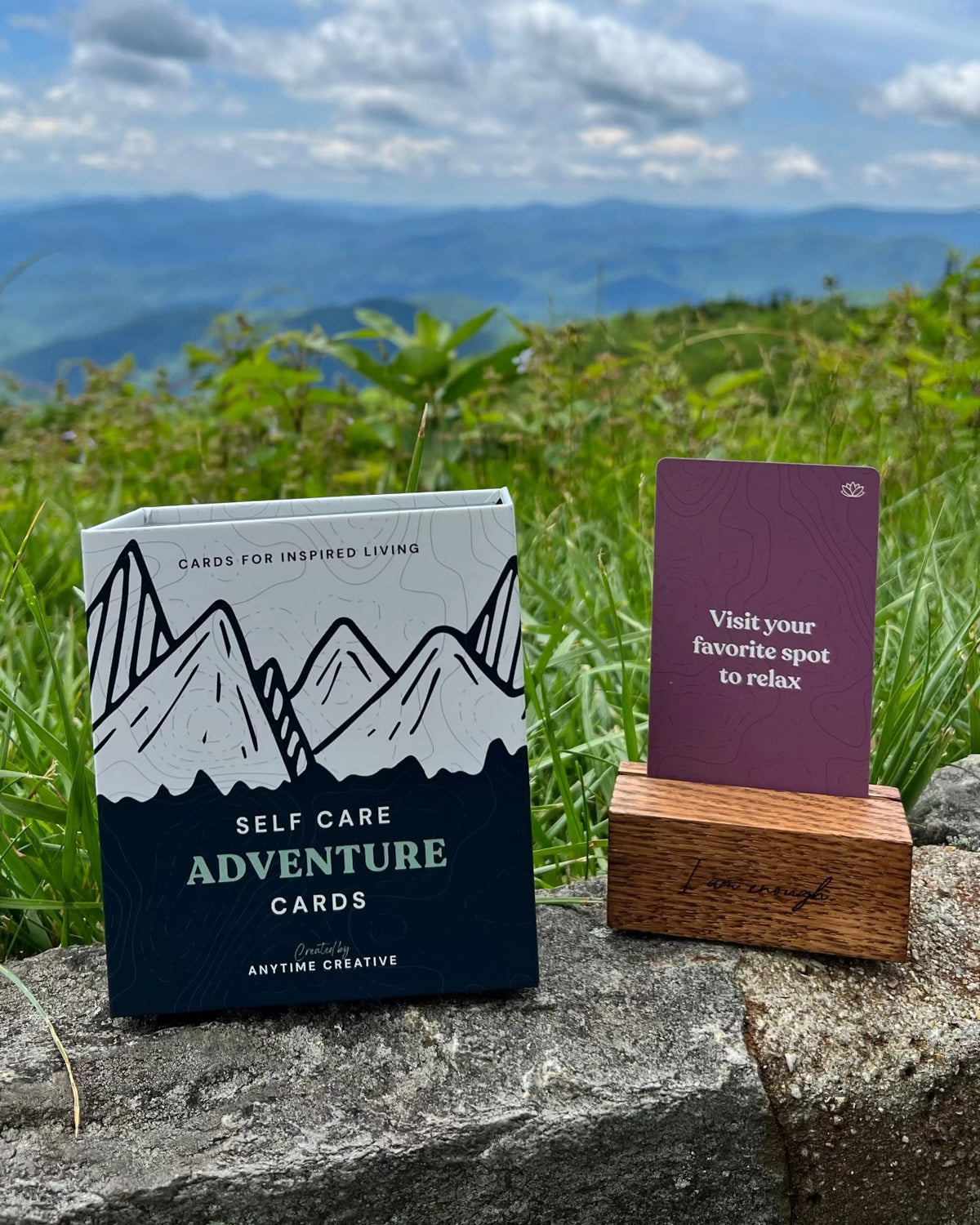 Self Care Adventure Cards box on a rock above green mountains, with a card in its stand