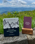 Self Care Adventure Cards box on a rock above green mountains, with a card in its stand