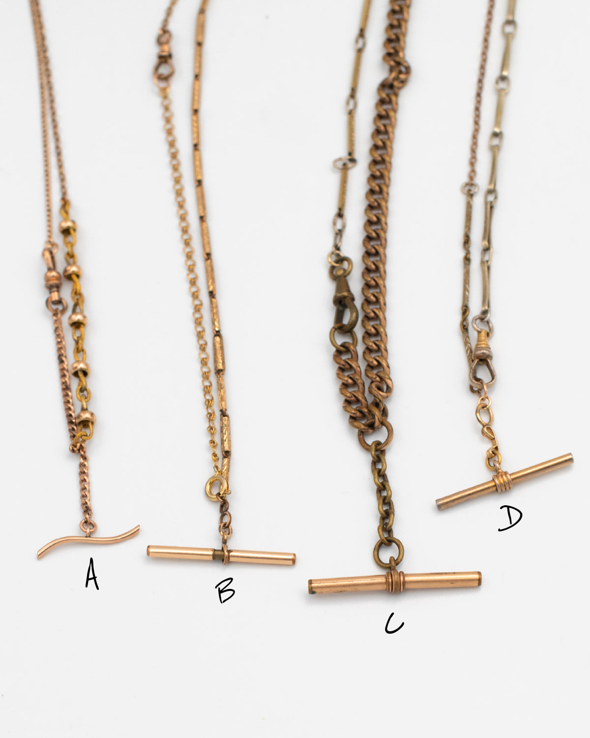 Close up of 4 necklaces labeled A, B, C, D made from Victorian-era men’s watch chains with antique T-bars and dog clips. Authentic 100+ year-old pieces made from sustainable vintage chains laying flat on a white background.