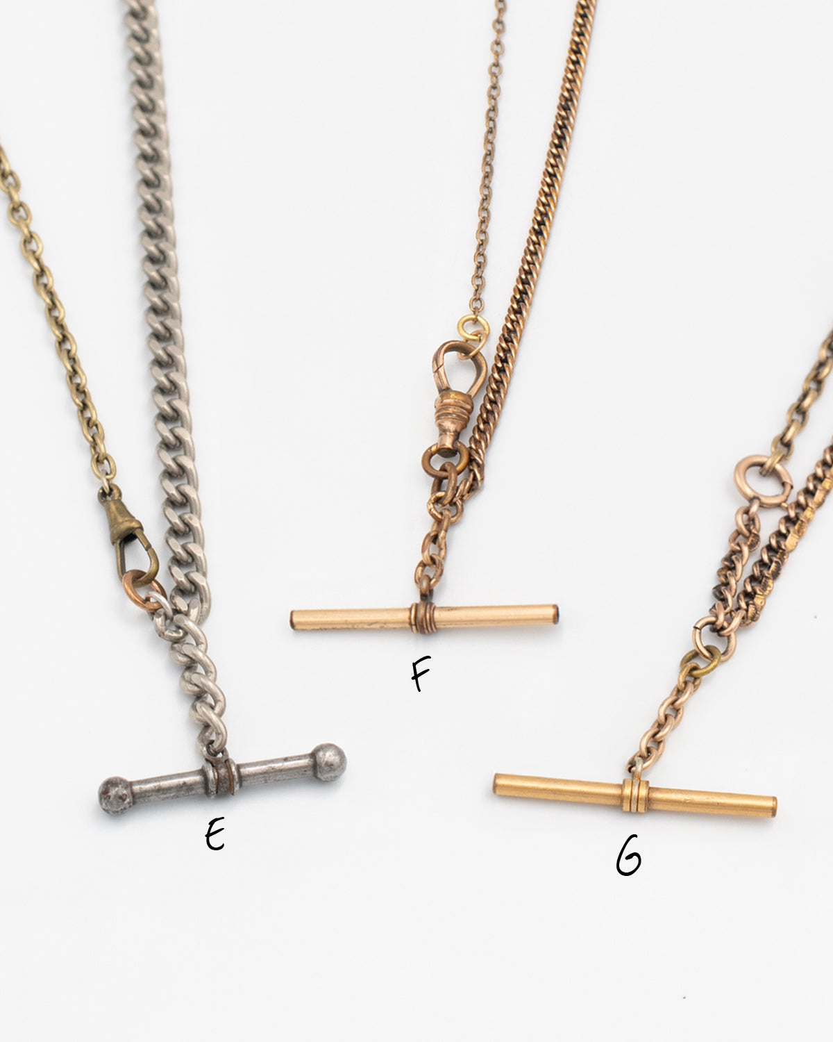 Close up of 3 necklaces labeled E, F, G made from Victorian-era men’s watch chains with antique T-bars and dog clips. Authentic 100+ year-old pieces made from sustainable vintage chains laying flat on a white background.