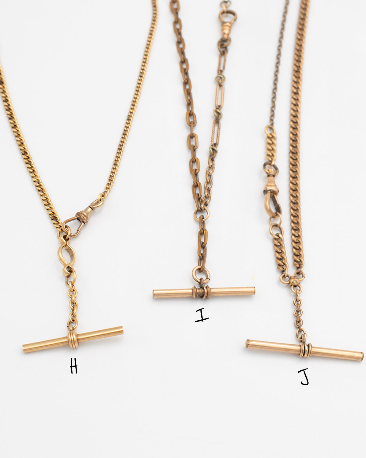 Close up of 4 necklaces labeled H, I, J made from Victorian-era men’s watch chains with antique T-bars and dog clips. Authentic 100+ year-old pieces made from sustainable vintage chains laying flat on a white background.
