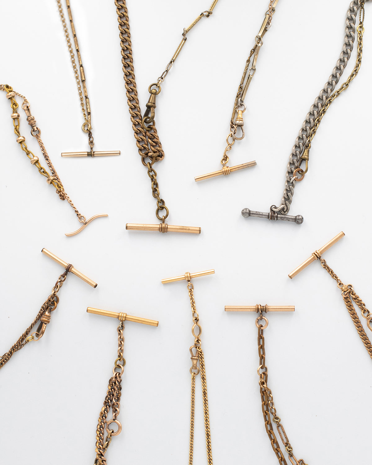 A collection of 10 necklaces made from Victorian-era men’s watch chains with antique T-bars and dog clips. Authentic 100+ year-old pieces made from sustainable vintage chains laying flat on a white background.