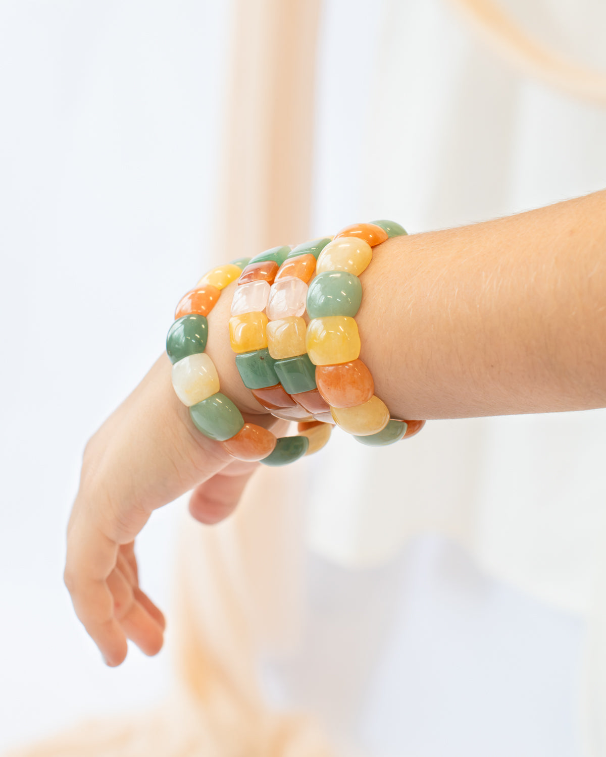 Woman's wrist stacked with multiple chicklet-style beaded agate stone bracelets in green, orange, yellow, and cream tones against a soft cream backdrop.