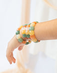Woman's wrist stacked with multiple chicklet-style beaded agate stone bracelets in green, orange, yellow, and cream tones against a soft cream backdrop.
