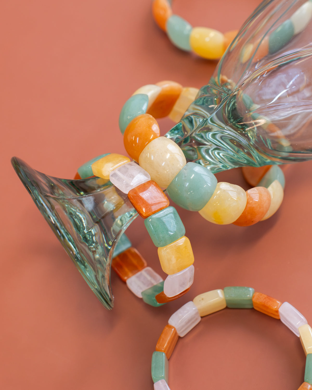 Chicklet beaded agate stone bracelets in green, orange, yellow, and cream draped over a glass vase against a terracotta background.