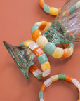 Chicklet beaded agate stone bracelets in green, orange, yellow, and cream draped over a glass vase against a terracotta background.