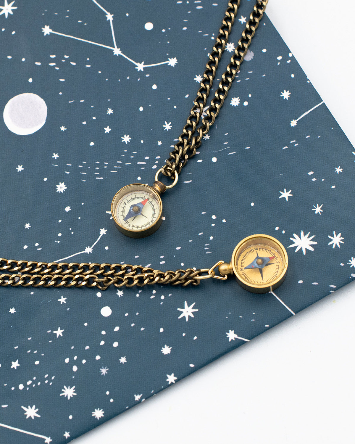 Two vintage compass necklaces on brass chains on a celestial themed background