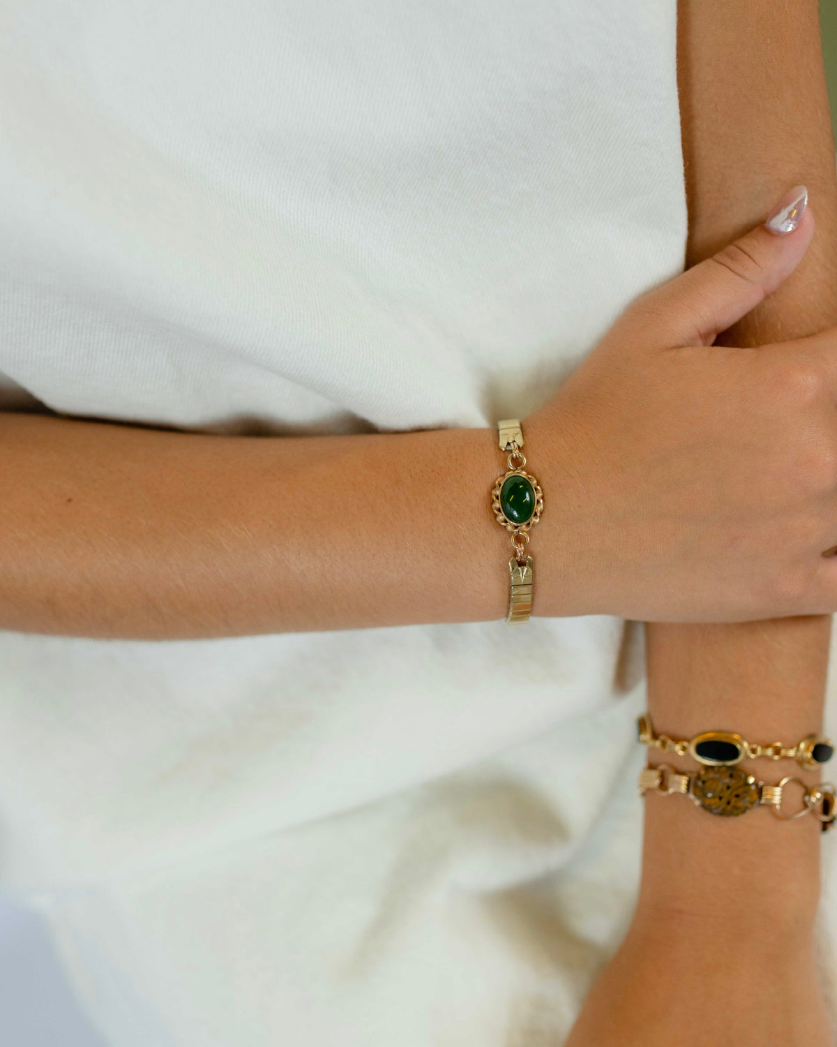 A close-up of a person's wrist wearing a gold-toned watchband bracelet with a prominent jade stone, paired with layered gold bracelets on another wrist, set against a soft white top.