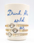 Stack of gold vintage bracelets on a ceramic holder with 'Drink the wild air' text.