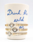 Stack of gold vintage bracelets on a ceramic holder with 'Drink the wild air' text.