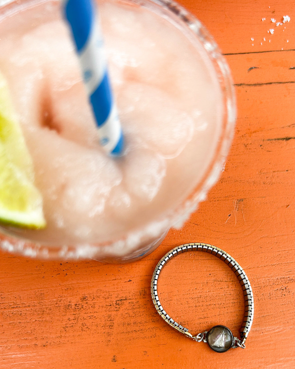 Close-up of a drink with a lime wedge and a straw, with a vintage hand-painted bracelet on an orange surface.