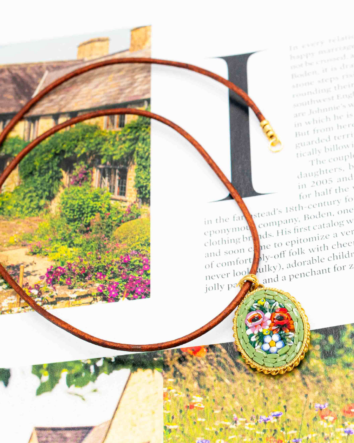 A brown leather cord necklace with a round, colorful floral micro mosaic pendant lies on top of an open book with a cottage and garden illustration.