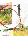 A brown leather cord necklace with a round, colorful floral micro mosaic pendant lies on top of an open book with a cottage and garden illustration.