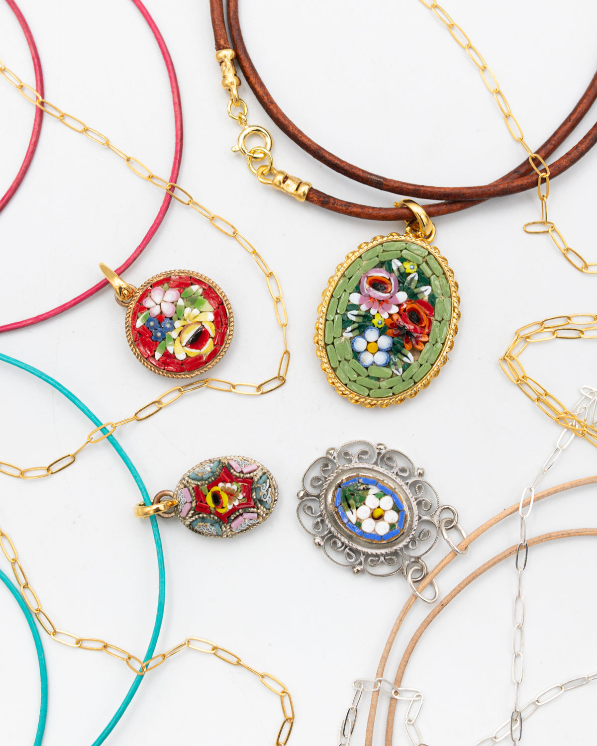 A styled flat-lay of several delicate micro mosaic necklaces on a white background, featuring colorful floral enamel and mosaic-style pendants in green, red, blue, and white, paired with gold, brown, pink, turquoise, and silver-tone chains.