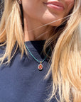 A close-up lifestyle shot of a woman wearing a turquoise cord necklace with a small shield-shaped micro mosaic pendant featuring a red and gold coat of arms.