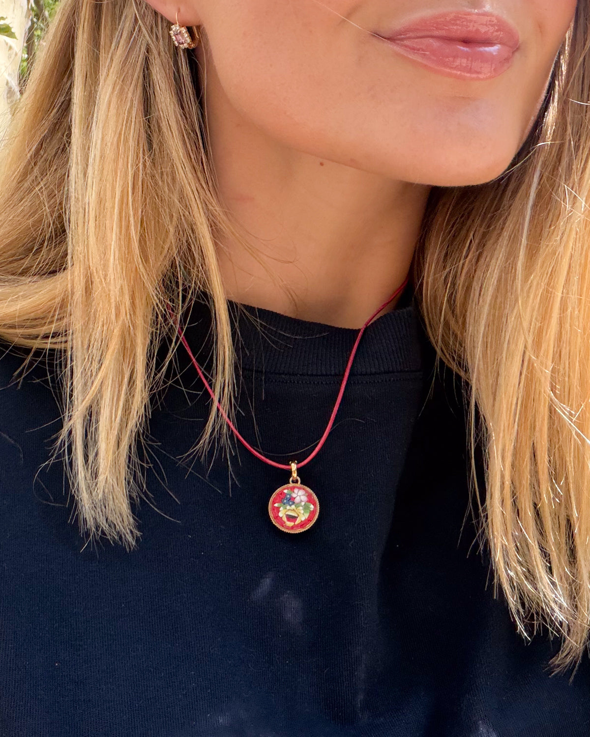 A close-up lifestyle shot of a woman wearing a red cord necklace with a round micro mosaic pendant featuring a colorful floral design and gold-toned metal.