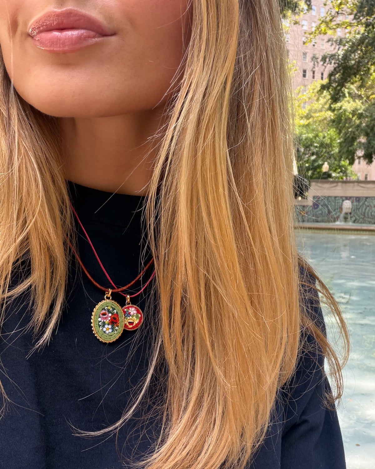 A close-up lifestyle shot of a woman with blonde hair wearing a double-strand red cord necklace with two oval micro mosaic pendant charms. One charm is gold-toned with a green background and red floral design, and the other is gold-toned with a red and green floral design.