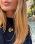 A close-up lifestyle shot of a woman with blonde hair wearing a double-strand red cord necklace with two oval micro mosaic pendant charms. One charm is gold-toned with a green background and red floral design, and the other is gold-toned with a red and green floral design.