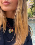A close-up lifestyle shot of a woman with blonde hair wearing a double-strand red cord necklace with two oval micro mosaic pendant charms. One charm is gold-toned with a green background and red floral design, and the other is gold-toned with a red and green floral design.