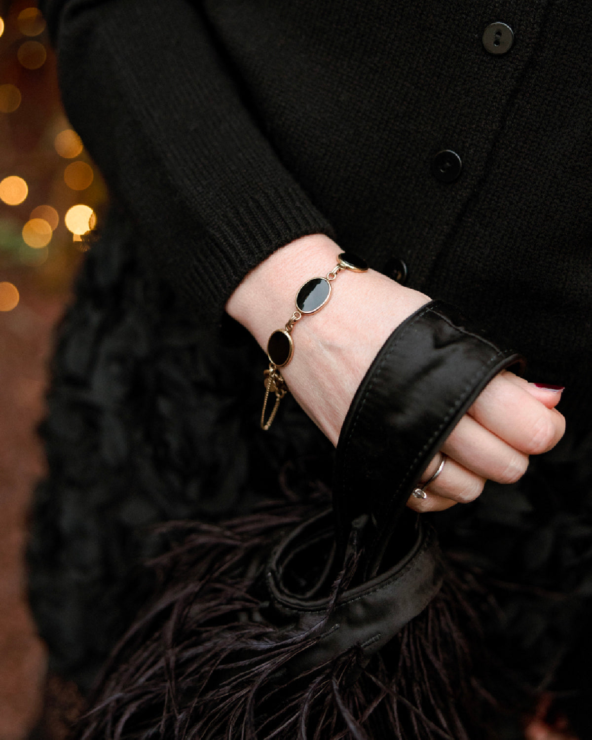 Close-up of a vintage gold-filled bracelet with black onyx cabochons worn with a black outfit and a feathered evening bag.