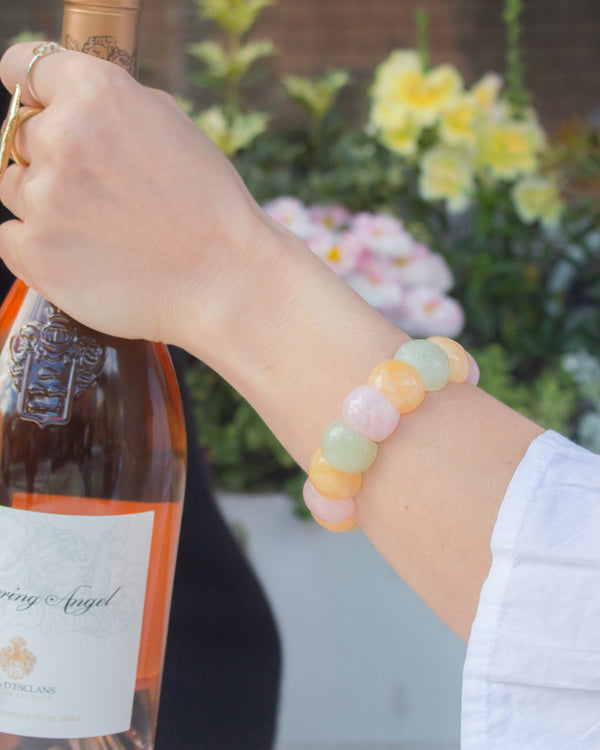 Woman wearing a pastel rainbow agate beaded bracelet in pink, yellow, and green tones while holding a bottle of rosé outdoors.