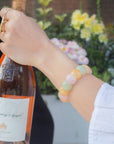 Woman wearing a pastel rainbow agate beaded bracelet in pink, yellow, and green tones while holding a bottle of rosé outdoors.