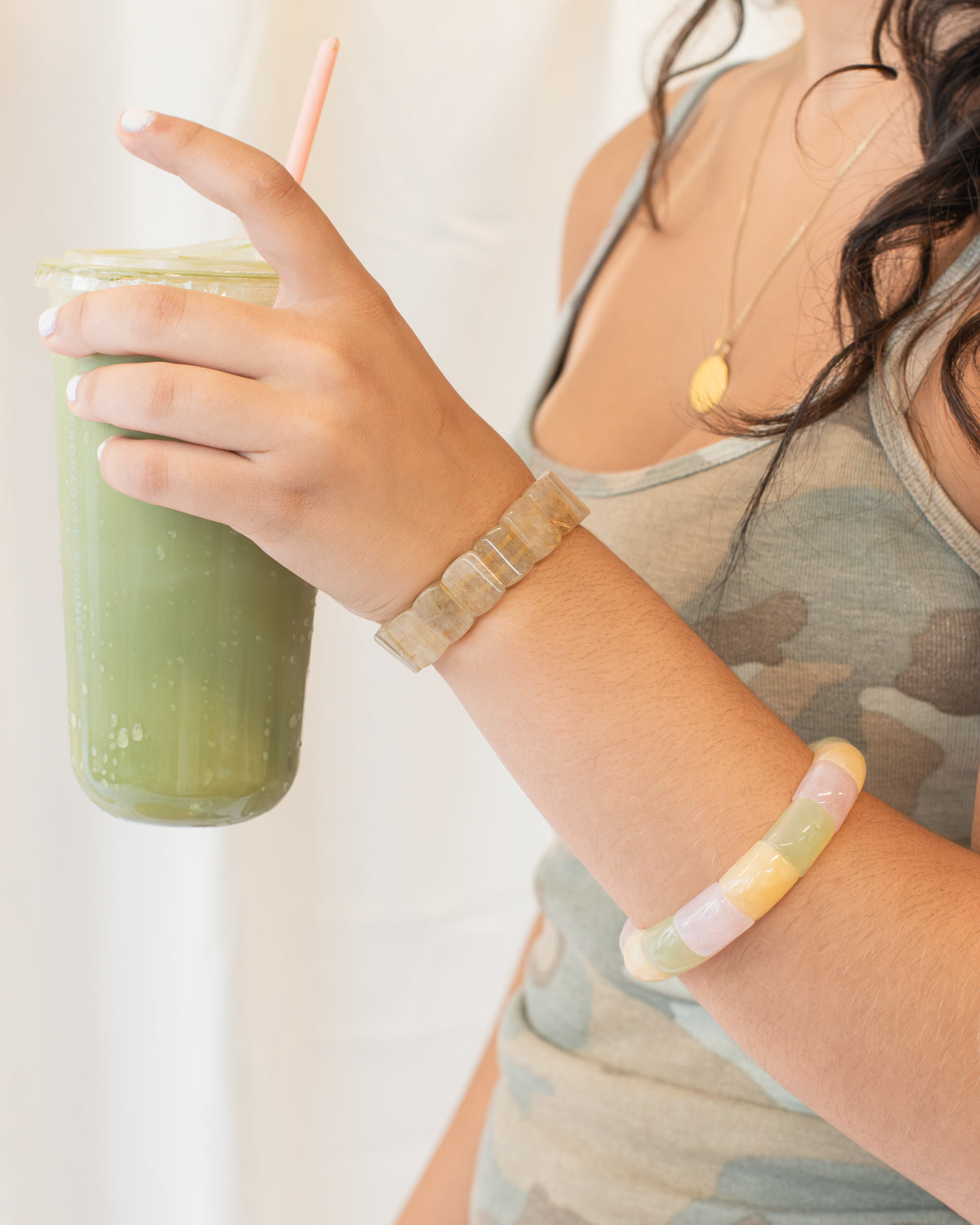 Woman holding a green iced drink while wearing a rutile quartz beaded bracelet stacked with a pink, yellow, and green agate bracelet.