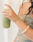 Woman holding a green iced drink while wearing a rutile quartz beaded bracelet stacked with a pink, yellow, and green agate bracelet.