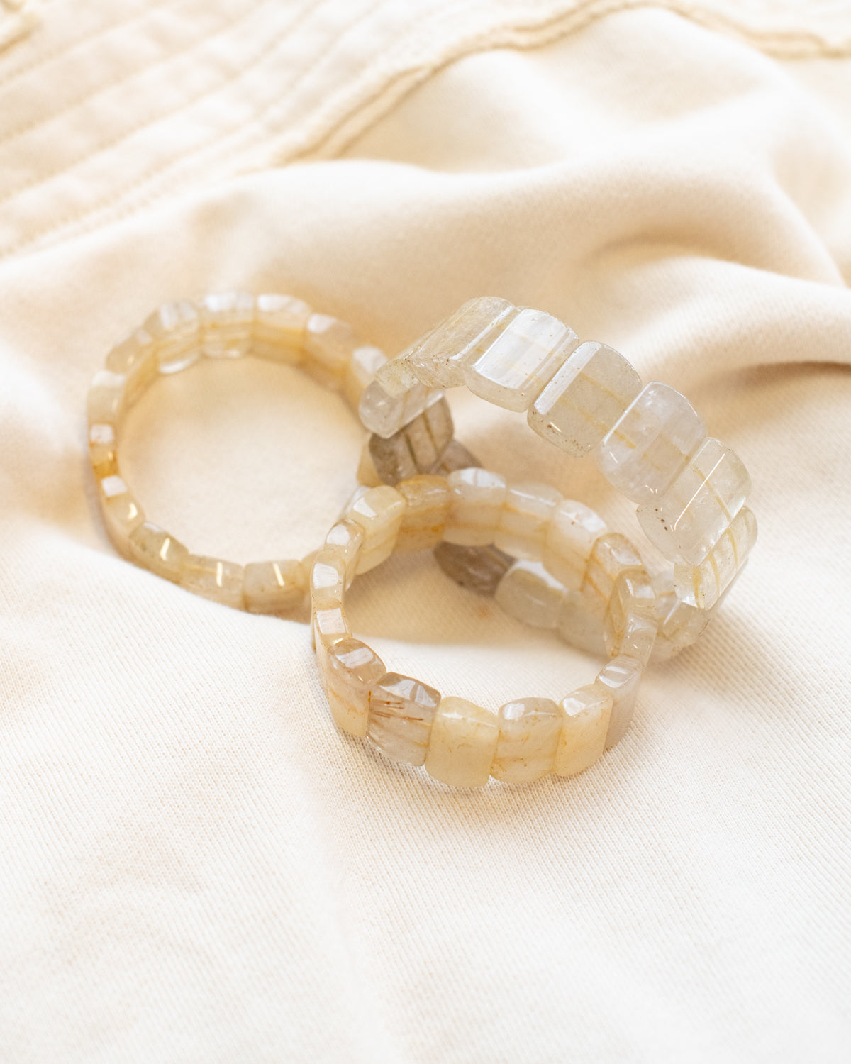Three rutile quartz beaded stone bracelets arranged on a cream fabric surface, showing their golden threaded inclusions.