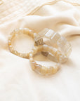 Three rutile quartz beaded stone bracelets arranged on a cream fabric surface, showing their golden threaded inclusions.