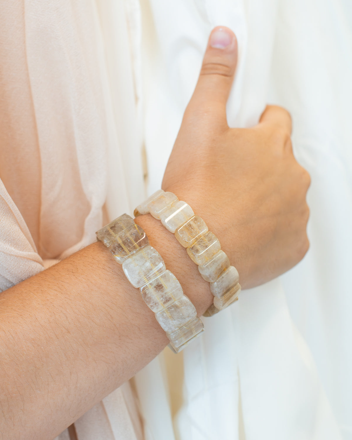 Close-up of two rutile quartz beaded bracelets stacked on a wrist against sheer cream fabric, highlighting their golden strands.