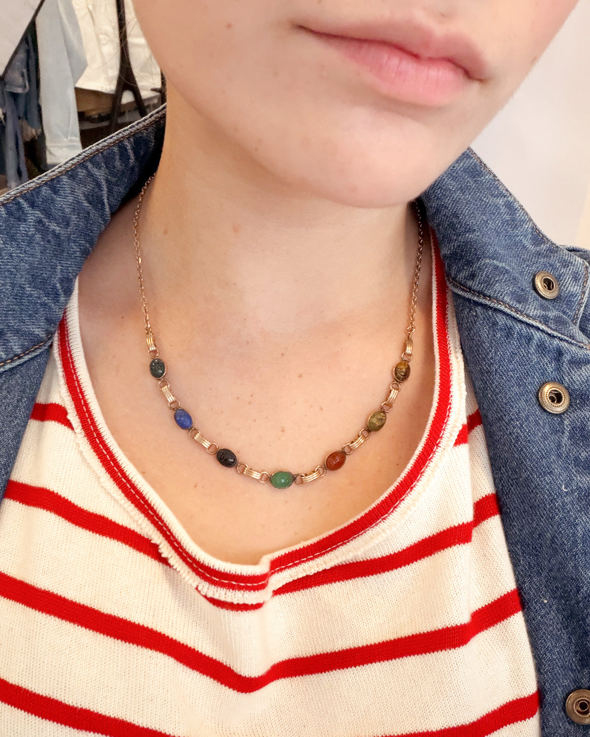 A close-up of a person wearing a delicate gold chain necklace with small multicolored beads, styled over a red and white striped shirt and a blue denim jacket.