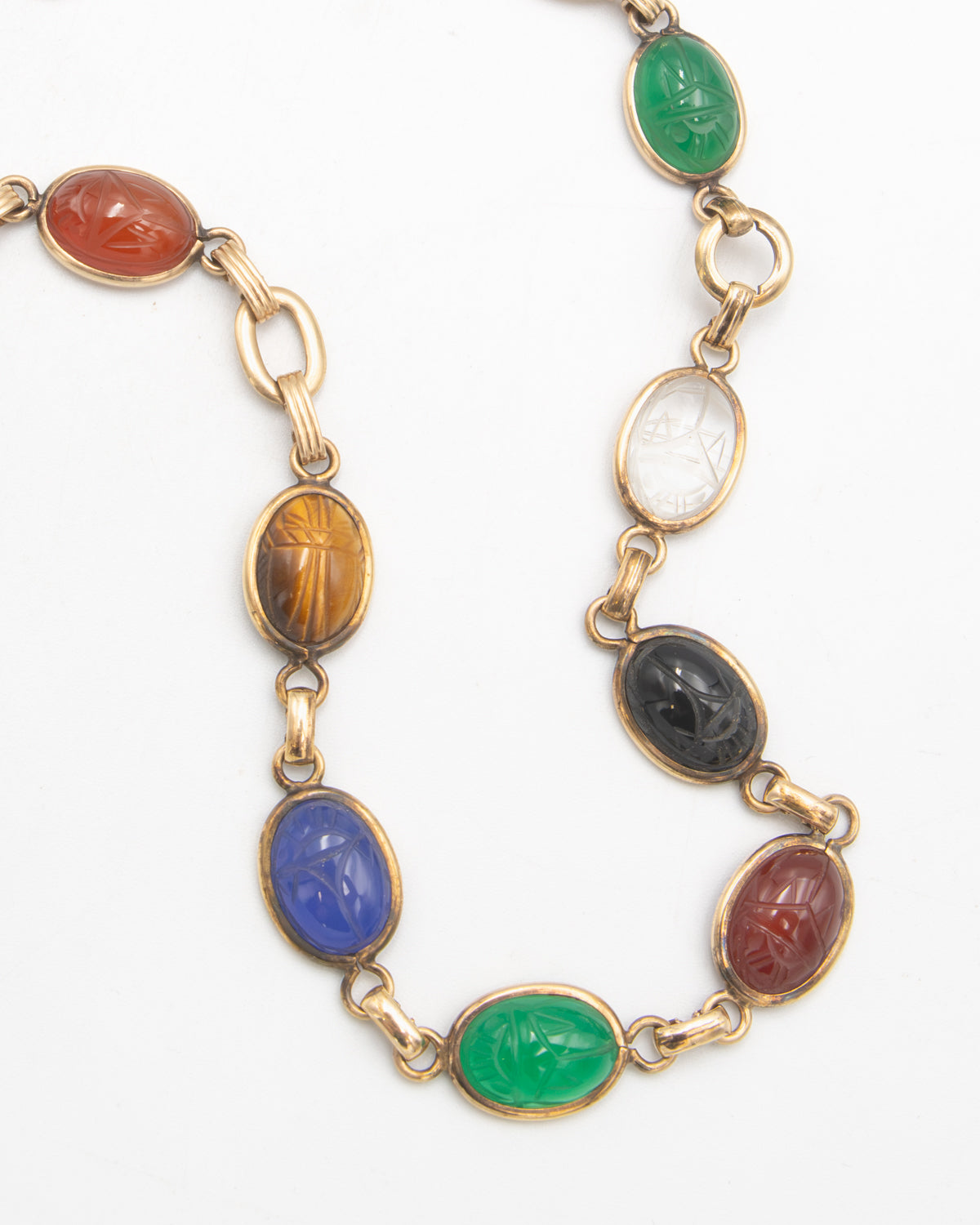 A gold-toned chain necklace featuring multiple oval cabochon gemstones carved with scarab beetle motifs in various colors including green, blue, brown, black, and red, set in gold links on a white background.