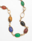 A gold-toned chain necklace featuring multiple oval cabochon gemstones carved with scarab beetle motifs in various colors including green, blue, brown, black, and red, set in gold links on a white background.