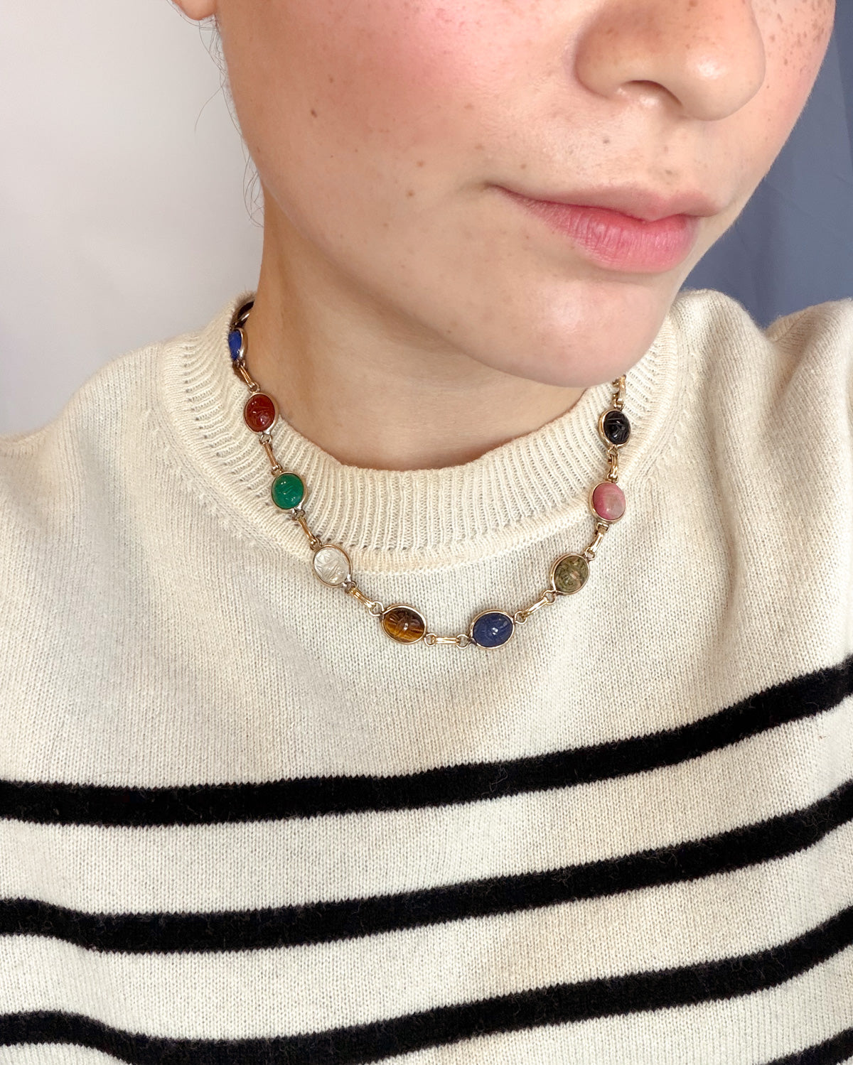 A close-up of a person wearing a vintage scarab gemstone necklace with colorful oval gemstone beads and gold-toned links, paired with a cream knit sweater with black horizontal stripes.