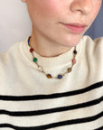 A close-up of a person wearing a vintage scarab gemstone necklace with colorful oval gemstone beads and gold-toned links, paired with a cream knit sweater with black horizontal stripes.