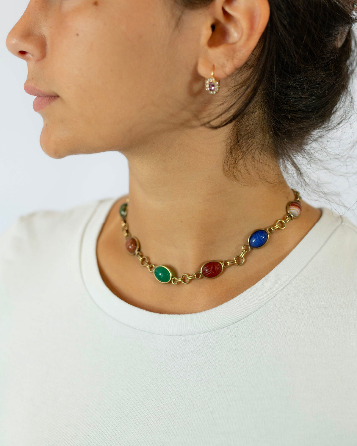 A close-up side profile of a woman wearing a gold-toned choker necklace with colorful oval cabochon scarab stones in green, red, and blue, paired with a white crewneck top.