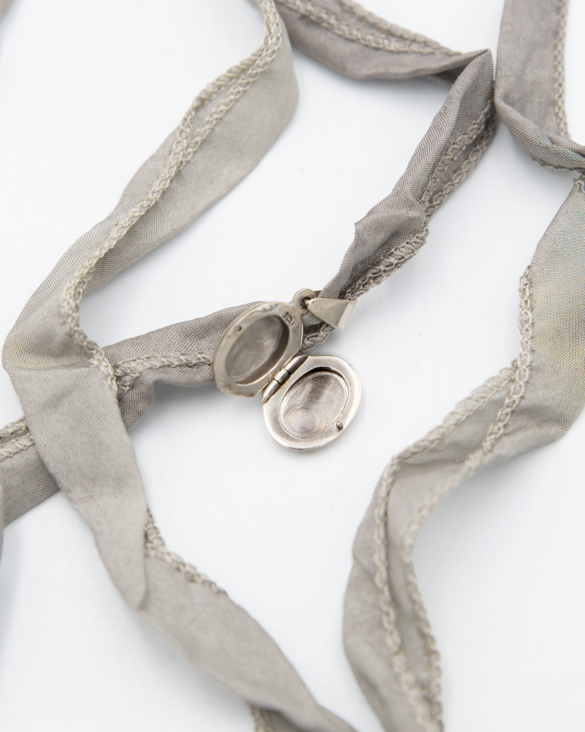 A silver oval locket pendant with a hinged split design, shown open on a white background, threaded onto a light gray silk ribbon with braided trim.