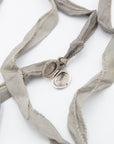 A silver oval locket pendant with a hinged split design, shown open on a white background, threaded onto a light gray silk ribbon with braided trim.