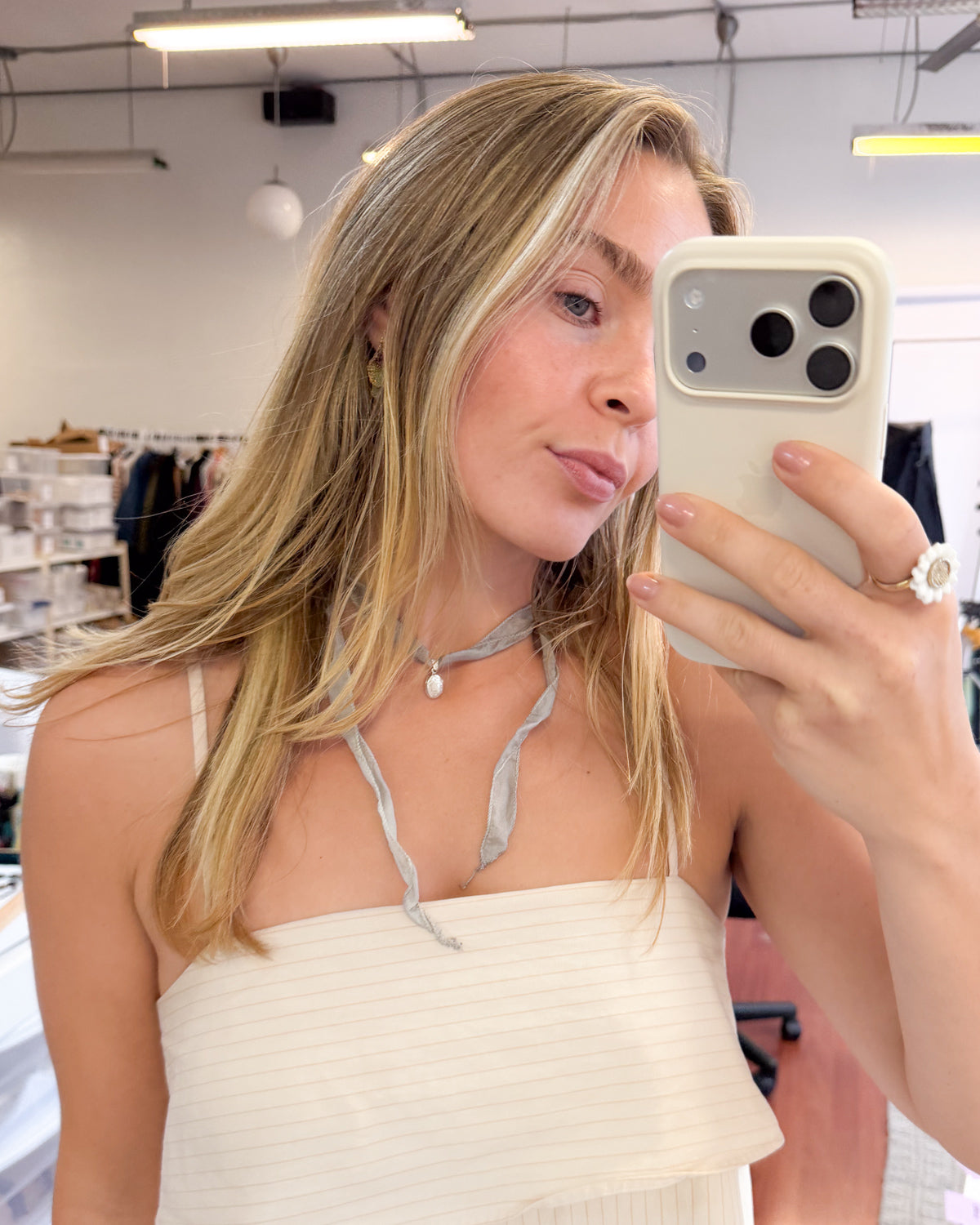 A woman taking a mirror selfie in a boutique, wearing a cream-colored ribbed tube top and a gray silk tie-necklace. She is holding a light beige smartphone.