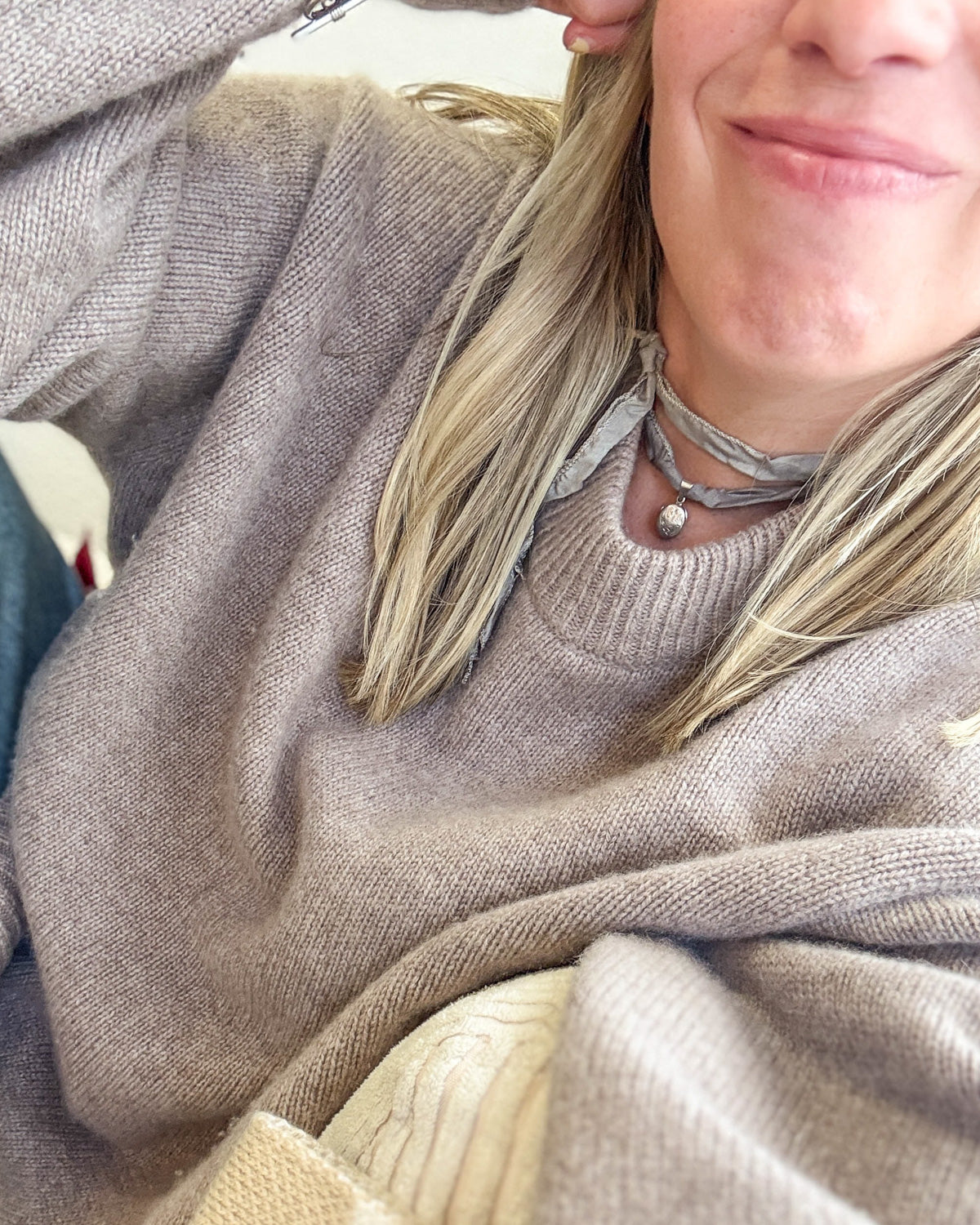 A close-up lifestyle shot of a person wearing a cozy, chunky knit crewneck sweater in a taupe-beige color. The person has blonde hair and is wearing a sterling silver vintage locket on a silk ribbon.