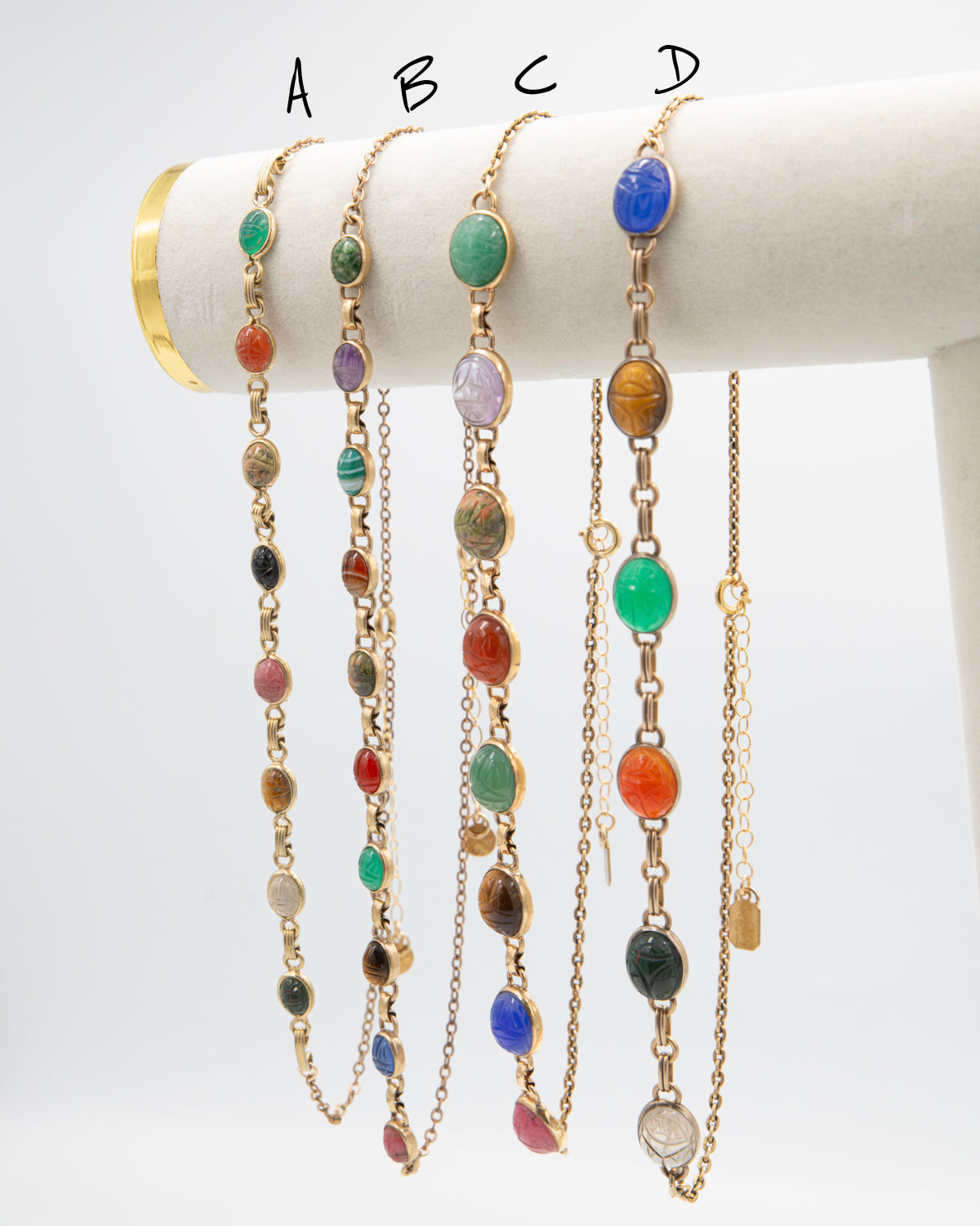 Four scarab beetle gemstone necklaces labeled A through D displayed side by side on a white jewelry stand.