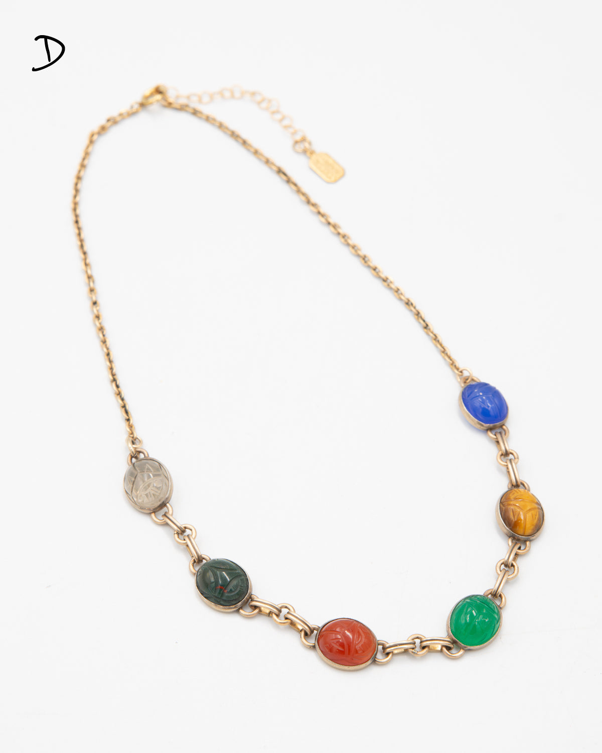 Scarab gemstone necklace featuring seven carved multicolor stone beetles linked in a gold chain, laid flat on a white background.