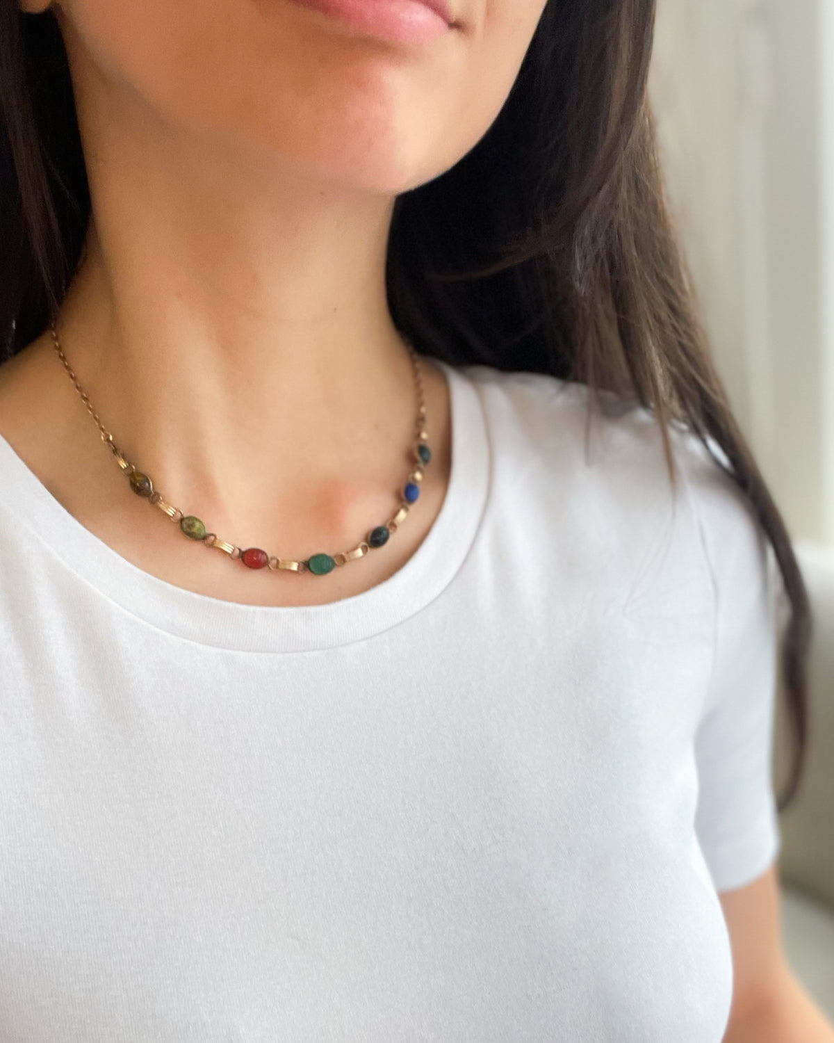 A close-up of a woman wearing a delicate gold chain necklace with small multicolored scarab gemstones, paired with a white crew-neck t-shirt.