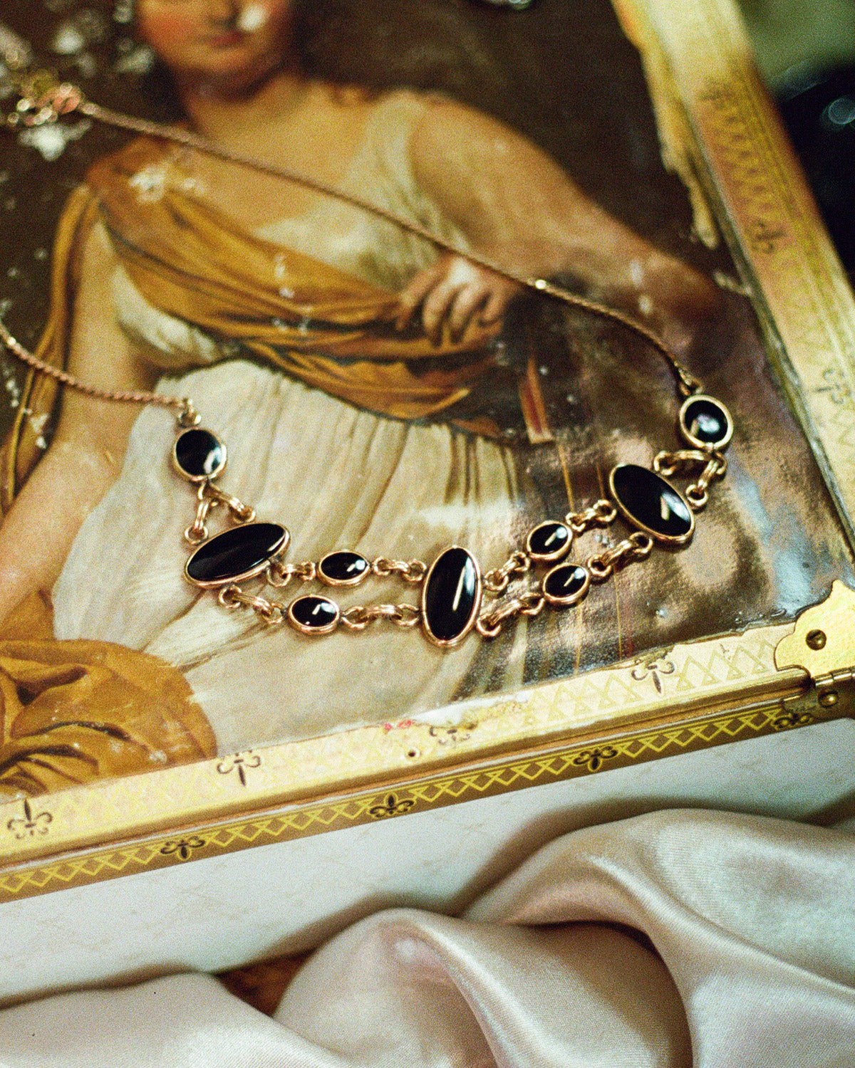 A gold-tone necklace with large and small oval black onyx stones, draped over an antique-style painting of a woman and resting on a white surface with satin fabric in the foreground.