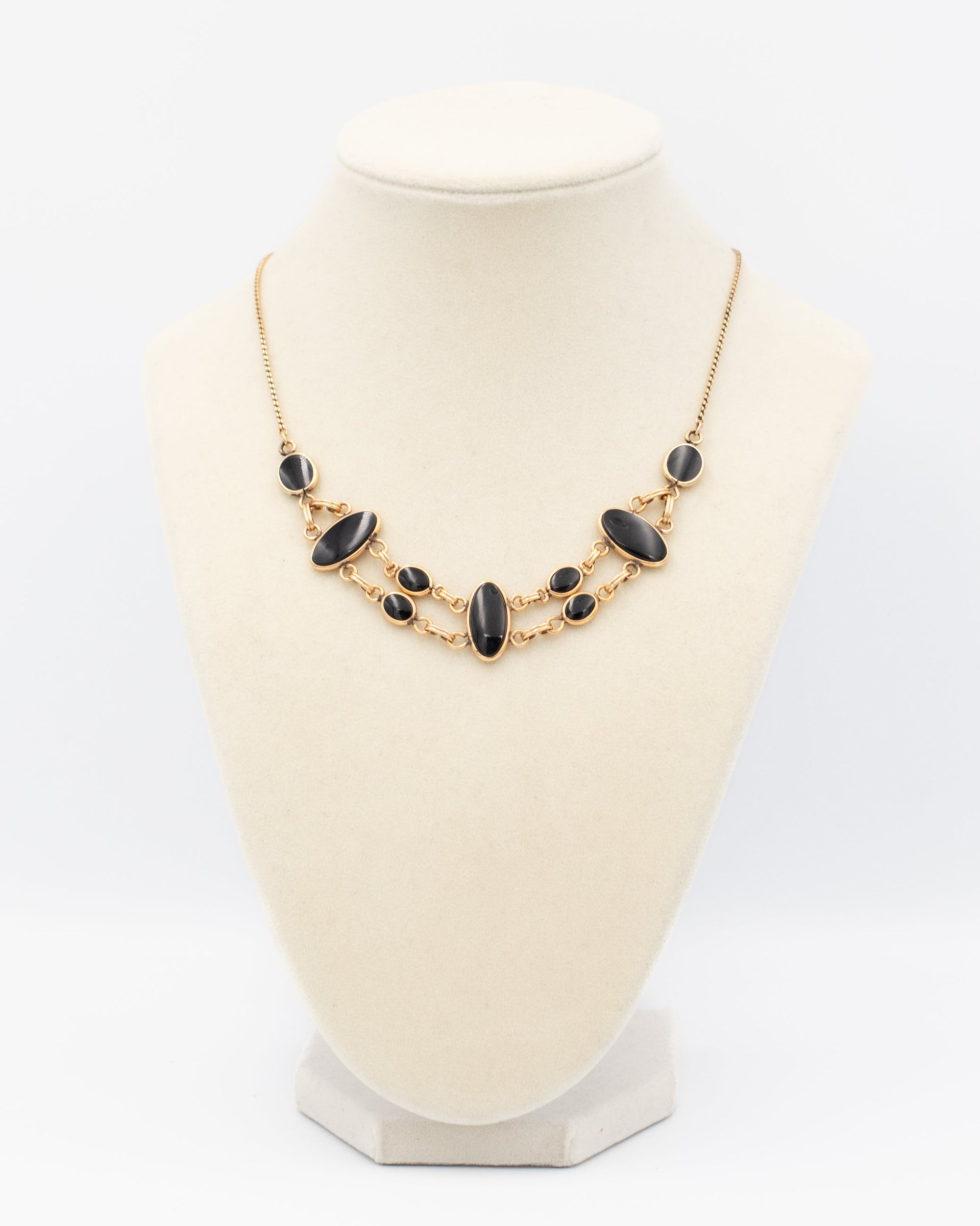 A gold statement necklace with dark oval and round onyx beads, displayed on a cream-colored jewelry bust against a white background.