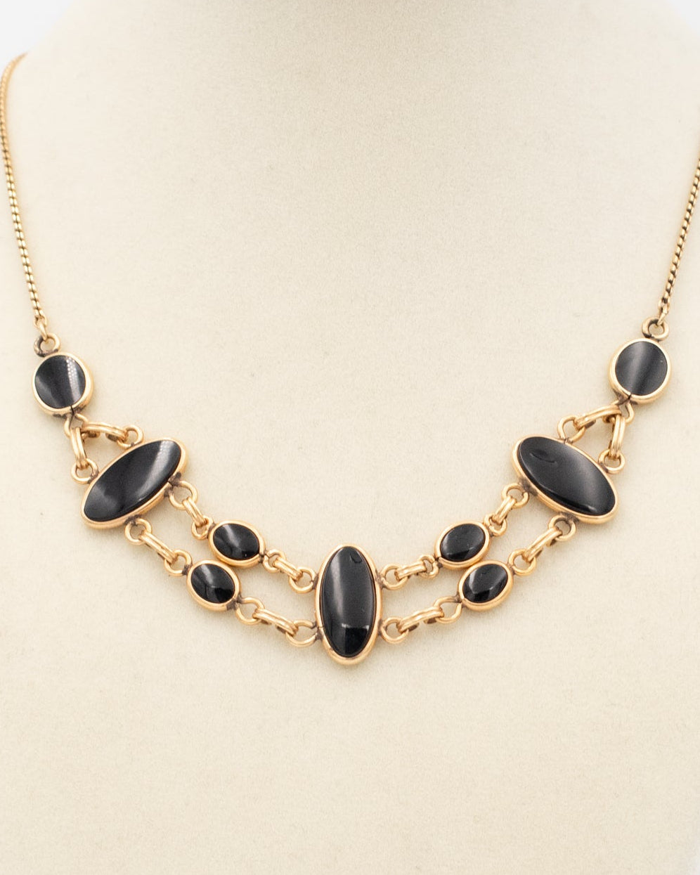 A gold statement necklace with dark oval and round onyx beads, displayed on a cream-colored jewelry bust against a white background.