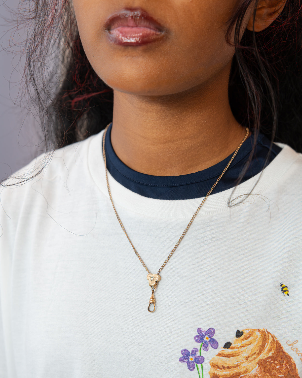 A close-up of a person wearing a delicate gold chain 19th century Victorian necklace with a small heart-shaped charm and a drop clasp, styled over a white ringer t-shirt with a navy collar.