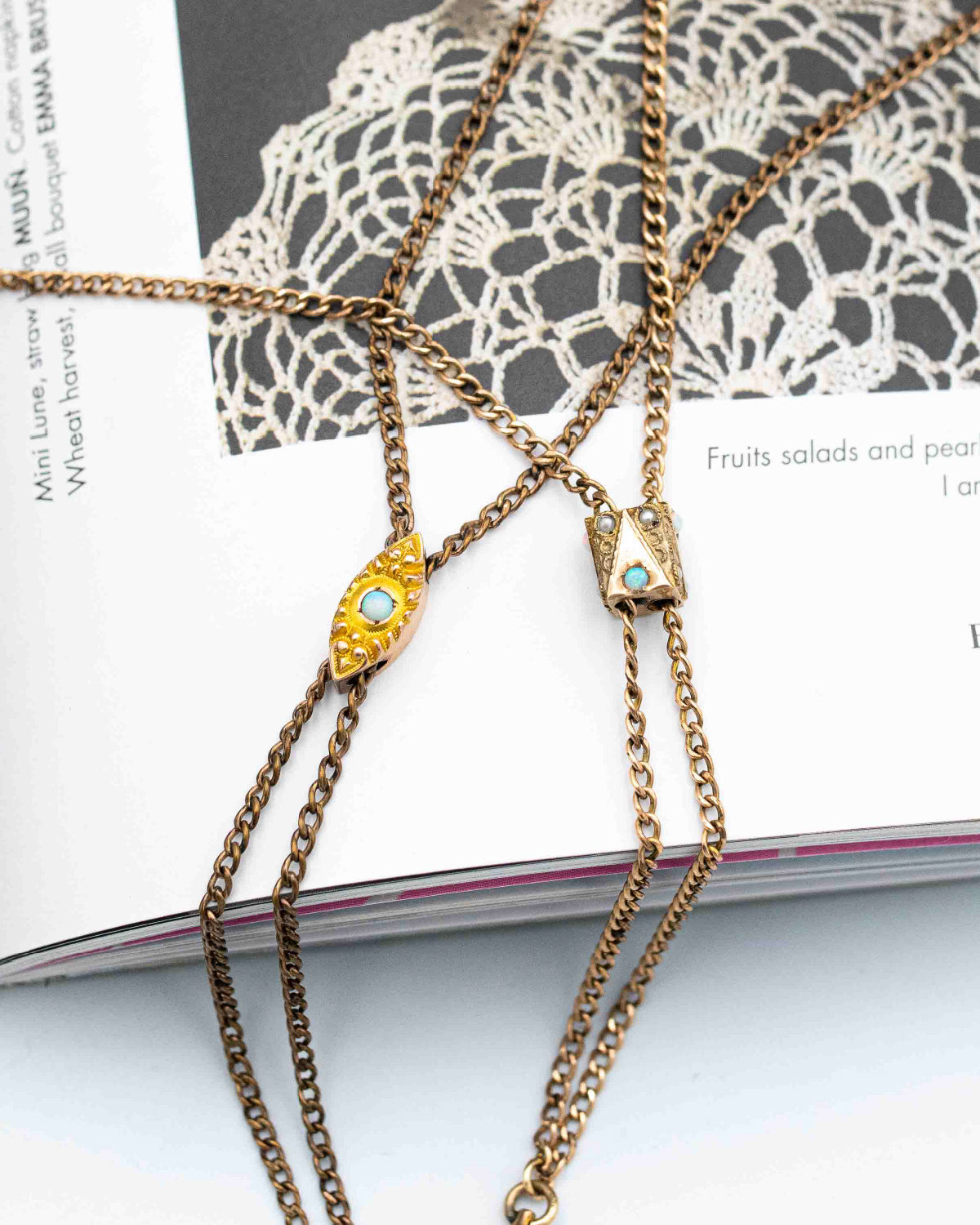 A close-up of layered gold-filled Victorian-era lariat slider chain necklaces draped over an open book. One pendant is an eye-shaped charm with a light blue opal stone, and another pendant has a small geometric box-like design with a matching opal blue stone.